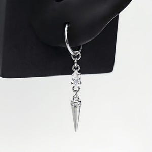 May include: A silver-toned hoop earring featuring a dangling charm. The charm consists of a small, clear marquise-cut gemstone connected to a metallic cone-shaped spike, which is accented with a row of tiny clear crystals at its base.