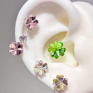 Op de afbeelding: Een verzameling bloemvormige oorstekers in verschillende kleuren, waaronder roze, groen en iriserend. De oorbellen worden gepresenteerd op een wit oor model, waarbij hun sprankelende, kristalachtige uiterlijk wordt benadrukt. De oorbellen zijn klein en delicaat.