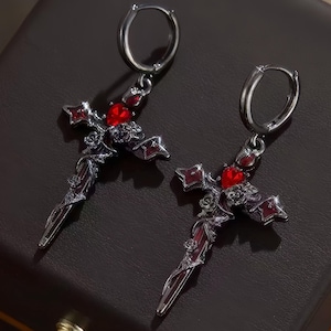 May include: A pair of silver-toned cross earrings with red gemstone accents. The earrings feature a detailed design with a dagger-like shape and a heart-shaped gem at the center of the cross. The earrings have a hoop closure.