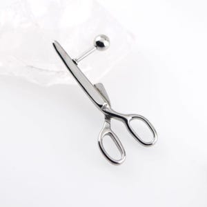 May include: A silver-toned metal body jewelry piece shaped like a pair of scissors. The design features a straight post with a small spherical ball end, attached to the scissor-shaped charm which has two oval finger loops and pointed blades.
