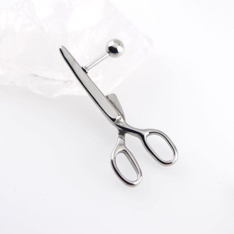 May include: A silver-toned metal body jewelry piece shaped like a pair of scissors. The design features a straight post with a small spherical ball end, attached to the scissor-shaped charm which has two oval finger loops and pointed blades.