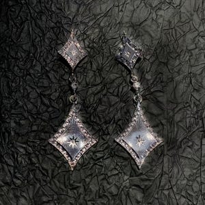 Puede incluir: Un par de pendientes colgantes plateados. Cada pendiente presenta un colgante en forma de diamante con un diseño de estrella y pequeños detalles brillantes. Los pendientes cuelgan de una pieza superior más pequeña, de forma cuadrada.