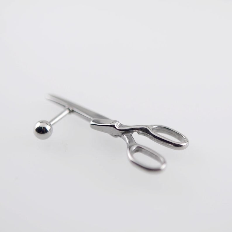 May include: A silver-toned metal body jewelry piece featuring a miniature scissor design. The jewelry includes a straight post with a small spherical ball end, connected to a detailed pair of scissors with two oval finger loops and blades.