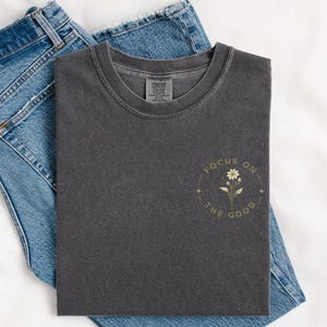May include: A charcoal gray short-sleeve t-shirt featuring a small graphic on the left chest. The graphic depicts a single flower surrounded by a circular border with the text "FOCUS ON THE GOOD" and small star accents. Blue denim jeans are underneath.
