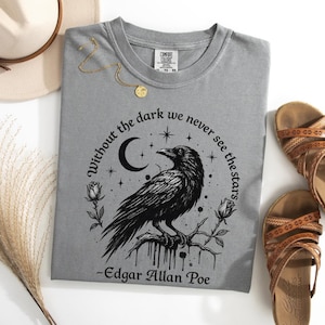 Op de afbeelding: Een grijs t-shirt met een zwarte afbeelding van een raaf op een tak, omringd door rozen, een maansikkel en sterren. De tekst luidt: "Without the dark we never see the stars - Edgar Allan Poe." Het shirt heeft een ronde halslijn.