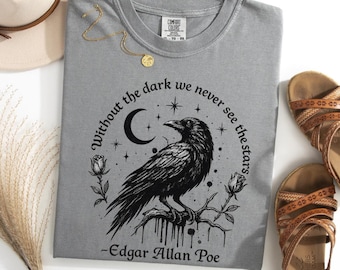 May include: A grey t-shirt featuring a black graphic of a raven perched on a branch, surrounded by roses, a crescent moon, and stars. The text reads, "Without the dark we never see the stars - Edgar Allan Poe." The shirt has a crew neckline.