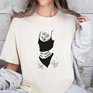 May include: A cream-colored short-sleeve t-shirt featuring a black graphic print of a torso silhouette with floral accents and the text "MY BODY, MY CHOICE" written in a bold, sans-serif font across the midsection of the design.
