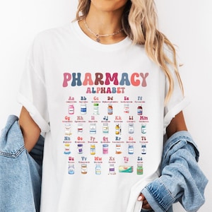 Op de afbeelding: Een wit T-shirt met korte mouwen en een kleurrijke afbeelding met de tekst PHARMACY ALPHABET. Het ontwerp toont de letters A tot en met Z, elk gecombineerd met een kleine illustratie van een medicijnflesje of medisch hulpmiddel in een raster.