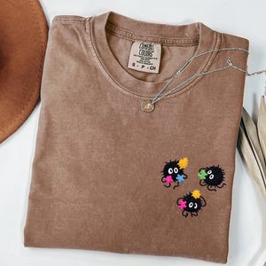 May include: A brown crewneck t-shirt with a Comfort Colors label in the collar. The shirt features an embroidered design on the lower right side consisting of three small, round, black fuzzy creatures with white eyes, each holding a colorful star.