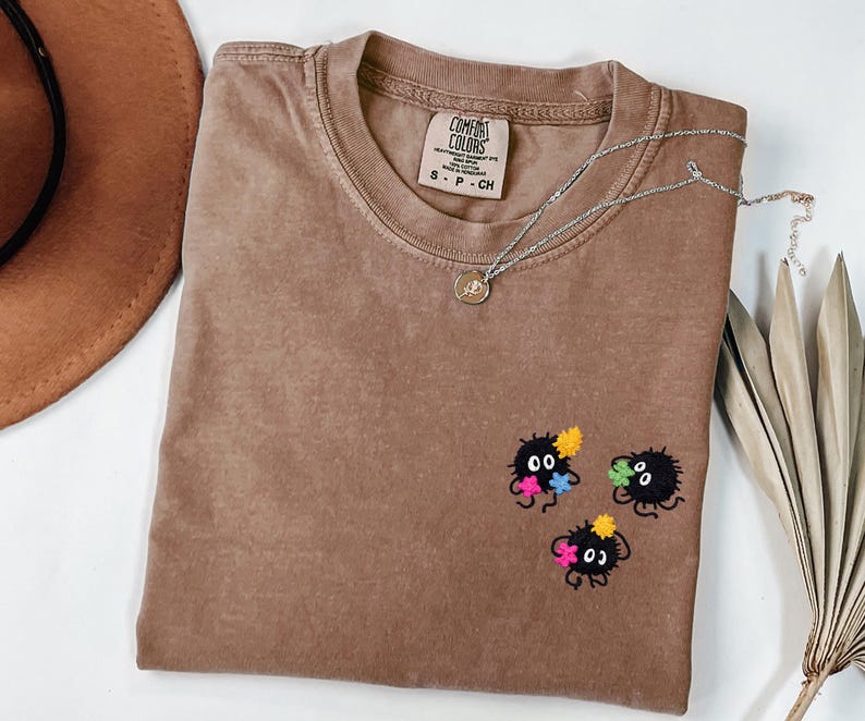 May include: A brown crewneck t-shirt with a Comfort Colors label in the collar. The shirt features an embroidered design on the lower right side consisting of three small, round, black fuzzy creatures with white eyes, each holding a colorful star.