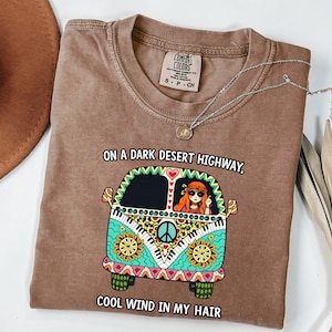 Op de afbeelding: Een bruin t-shirt met korte mouwen en een afbeelding van een kleurrijk busje met een persoon erin. De tekst luidt: "ON A DARK DESERT HIGHWAY, COOL WIND IN MY HAIR." Het ontwerp van het busje bevat patronen van bloemen, vredestekens en pianotoetsen.