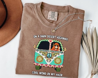 May include: A brown short-sleeve t-shirt featuring a graphic of a colorful van with a person inside. The text reads, "ON A DARK DESERT HIGHWAY, COOL WIND IN MY HAIR." The van design includes patterns of flowers, peace signs, and piano keys in bright colors.