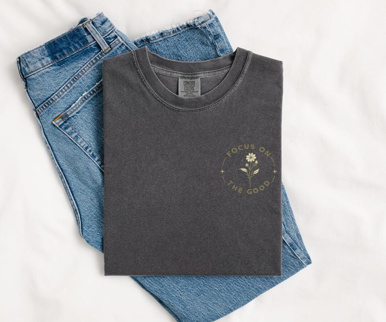 May include: A charcoal gray short-sleeve t-shirt featuring a small graphic on the left chest. The graphic depicts a single flower surrounded by a circular border with the text "FOCUS ON THE GOOD" and small star accents. Blue denim jeans are underneath.