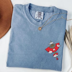 Op de afbeelding: Een blauw T-shirt met korte mouwen en een Comfort Colors-label in de nek. Het shirt heeft een geborduurde afbeelding aan de rechteronderkant van een rode papieren paraplu met gouden baleinen en witte wolken, afgewerkt met een rode kwast.