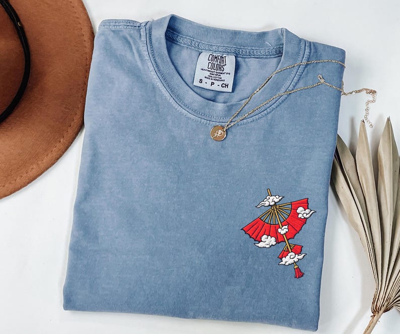 Op de afbeelding: Een blauw T-shirt met korte mouwen en een Comfort Colors-label in de nek. Het shirt heeft een geborduurde afbeelding aan de rechteronderkant van een rode papieren paraplu met gouden baleinen en witte wolken, afgewerkt met een rode kwast.