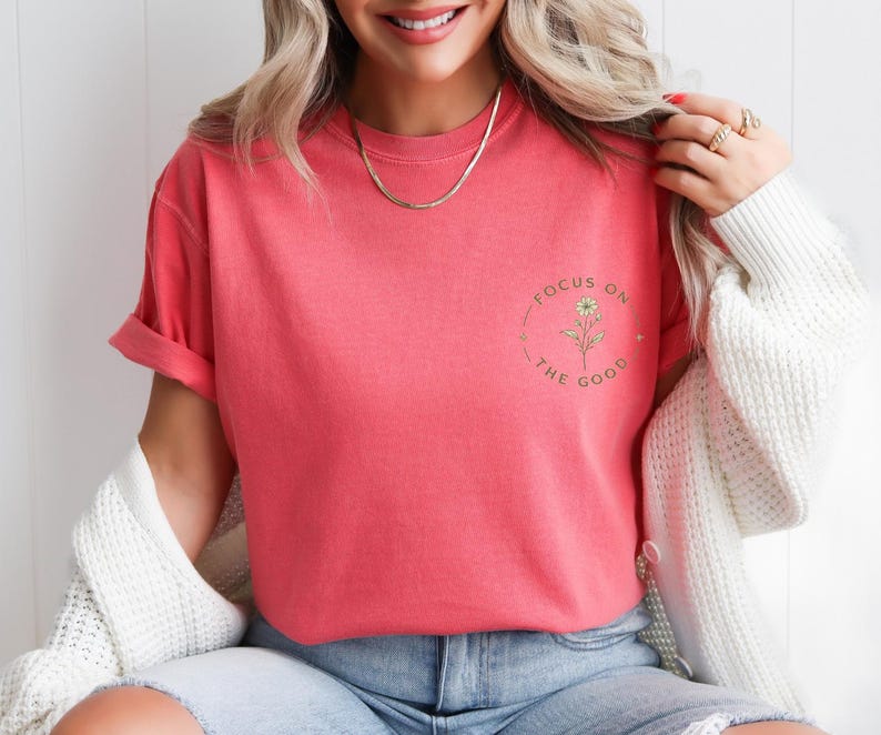 May include: A coral-colored short-sleeve t-shirt featuring a small circular graphic on the left chest. The graphic contains a line-art flower illustration surrounded by the text "FOCUS ON THE GOOD" in a simple, dark-colored sans-serif font.
