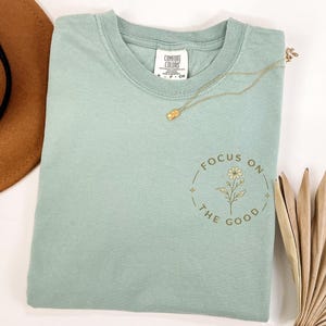 May include: A sage green short-sleeved t-shirt featuring a Comfort Colors brand label at the inner neckline. The front of the shirt displays a circular graphic with a small flower illustration and the text "FOCUS ON THE GOOD" in gold.