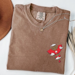 Op de afbeelding: Een bruin t-shirt met korte mouwen, een ronde hals en een Comfort Colors merklabel. Het shirt heeft een geborduurde afbeelding aan de rechteronderkant met een rode parasol met witte wolken en een klein kwastje dat onderaan hangt.