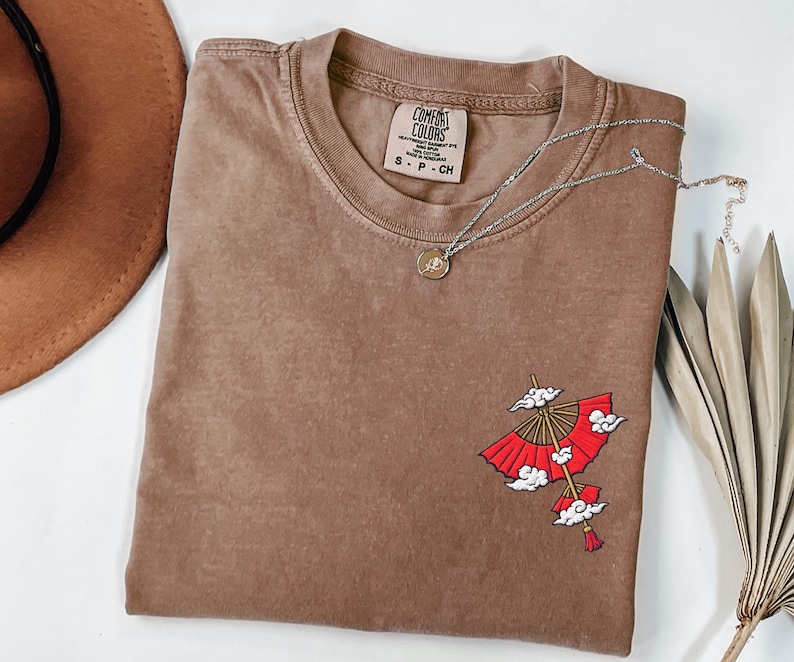 Op de afbeelding: Een bruin t-shirt met korte mouwen, een ronde hals en een Comfort Colors merklabel. Het shirt heeft een geborduurde afbeelding aan de rechteronderkant met een rode parasol met witte wolken en een klein kwastje dat onderaan hangt.