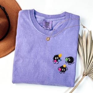 May include: A lavender short-sleeved t-shirt with a Comfort Colors label at the neck. The shirt features three small, black, fuzzy, circular creatures embroidered on the chest, each holding tiny, colorful, star-shaped flowers in yellow, pink, and blue.