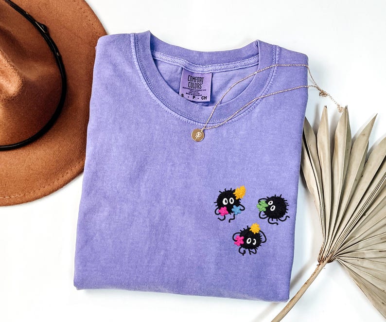 May include: A lavender short-sleeved t-shirt with a Comfort Colors label at the neck. The shirt features three small, black, fuzzy, circular creatures embroidered on the chest, each holding tiny, colorful, star-shaped flowers in yellow, pink, and blue.