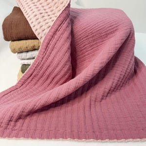 Peut inclure: Une pile de couvertures pliées de différentes couleurs, dont marron, beige, gris et vert olive. Une grande couverture rose poussiéreux est drapée sur la pile, avec une section texturée rose clair en haut. Les couvertures ont une texture douce et froissée.