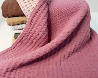 May include: A stack of folded blankets in various colors, including brown, tan, gray, and olive green. A large, dusty rose-colored blanket is draped over the stack, with a textured, light pink section at the top. The blankets have a soft, crinkled texture.