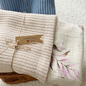 May include: A beige crocheted blanket is tied with twine and a small tag that reads "Handmade with love." A floral patterned blanket is visible next to the crocheted blanket. A blue crocheted blanket is in the background. The blankets are in a woven basket.