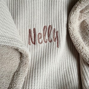 May include: Close-up of a beige knitted blanket with the name "Nelly" embroidered in a brown script font. The blanket has a soft, plush lining in a lighter beige color, providing a cozy texture.
