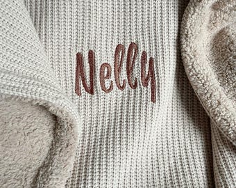 May include: Close-up of a beige knitted blanket with the name "Nelly" embroidered in a brown script font. The blanket has a soft, plush lining in a lighter beige color, providing a cozy texture.