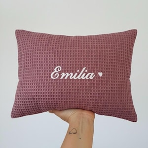 May include: A mauve rectangular cushion with a waffle-weave texture. The name "Emilia" is embroidered in white script across the cushion, with a small white heart beside it.
