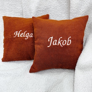 May include: Two square, rust-colored, plush-textured throw pillows. The left pillow features the white embroidered name "Helga" in a cursive script, while the right pillow displays the white embroidered name "Jakob" in a matching cursive script.