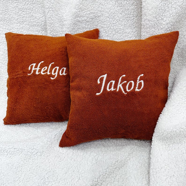 May include: Two square, rust-colored, plush-textured throw pillows. The left pillow features the white embroidered name "Helga" in a cursive script, while the right pillow displays the white embroidered name "Jakob" in a matching cursive script.