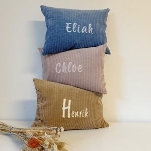 May include: Three corduroy pillows stacked vertically. The top pillow is blue with the name "Eliah" embroidered in white. The middle pillow is pink with "Chloe" embroidered in white. The bottom pillow is tan with "Henrik" embroidered in white.