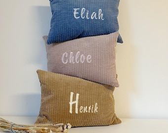 May include: Three corduroy pillows stacked vertically. The top pillow is blue with the name "Eliah" embroidered in white. The middle pillow is pink with "Chloe" embroidered in white. The bottom pillow is tan with "Henrik" embroidered in white.