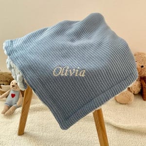 May include: A light blue knitted baby blanket with the name "Olivia" embroidered in white. The blanket is folded over a small wooden stool. A teddy bear and a stuffed toy are in the background.