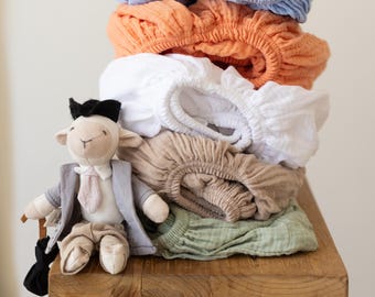 May include: A stack of colorful fitted sheets in blue, orange, white, tan, and green, resting on a wooden bench. A stuffed lamb wearing a suit and holding an umbrella sits on the bench next to the sheets.