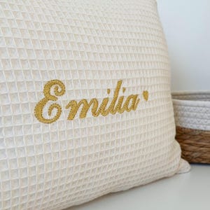 A highly detailed close-up shot of a cream-colored waffle-weave cushion, showcasing the intricate and high-quality artisan embroidery of the name "Emilia" in white script thread, resting on a rustic wooden workbench.

Eine sehr detaillierte Nahaufnahme eines cremefarbenen Kissens im Waffelpikee-Muster, die die aufwendige und hochwertige handwerkliche Stickerei des Namens "Emilia" in weißem Garn auf einer rustikalen Holzwerkbank zeigt.