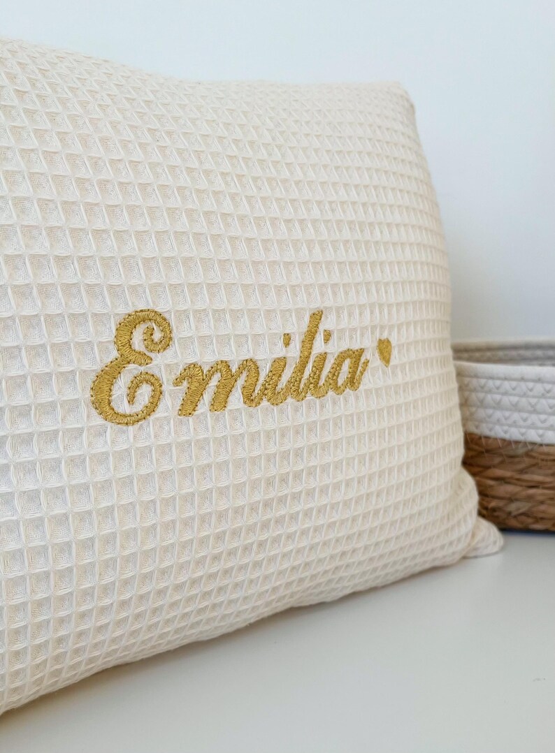 A highly detailed close-up shot of a cream-colored waffle-weave cushion, showcasing the intricate and high-quality artisan embroidery of the name "Emilia" in white script thread, resting on a rustic wooden workbench.

Eine sehr detaillierte Nahaufnahme eines cremefarbenen Kissens im Waffelpikee-Muster, die die aufwendige und hochwertige handwerkliche Stickerei des Namens "Emilia" in weißem Garn auf einer rustikalen Holzwerkbank zeigt.