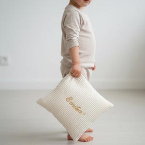Könnte beinhalten: Ein cremefarbenes, quadratisches Kissen mit Waffelstruktur und dem Namen "Emilia" in goldfarbenem Faden gestickt. Das Kissen wird von einem Kind gehalten, das ein beiges Langarmshirt und eine passende Hose trägt.