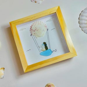 May include: A framed artwork featuring a seashell hot air balloon design. The balloon is a large, white and pink seashell, with a blue glass swing below. The frame is a light yellow color. Seashells are scattered around the frame.