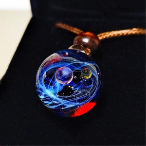May include: A glass pendant necklace with a miniature galaxy design. The pendant is a sphere with swirling blue, white, and red colours, and includes small, colourful orbs. The necklace has a brown cord.