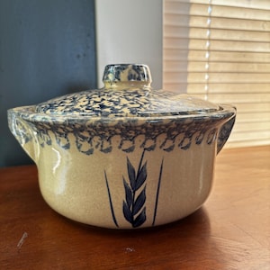 May include: A ceramic casserole dish with a lid. The dish is beige with blue speckled patterns and a blue wheat stalk design. The lid has a knob and matches the dish's pattern. The dish has two handles.