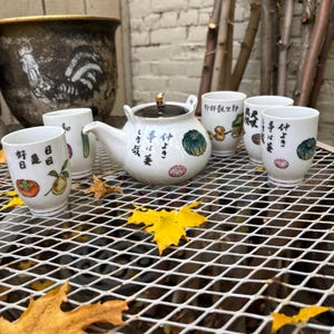 May include: A white ceramic tea set with a teapot and six matching cups. The teapot has a black lid and gold accents. The cups and teapot are decorated with Japanese characters and colorful illustrations of fruits and vegetables.
