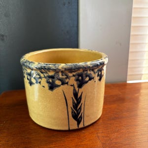 May include: A cylindrical, yellow ceramic container with a blue floral and wheat design. The rim features a blue floral pattern, and a wheat stalk is painted on the side. The container sits on a wooden surface.