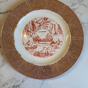 May include: A vintage decorative plate with a pink and gold rim featuring an intricate design. The centre of the plate displays a red and white illustration of Yosemite National Park, with text that reads "Yosemite Natl. Park California".