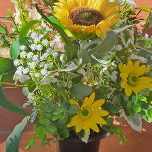 May include: A floral arrangement in a black pot featuring a large yellow sunflower, smaller yellow daisies, white baby's breath, and various green eucalyptus leaves. The bouquet is dense with foliage and flowers, creating a full, rounded display.