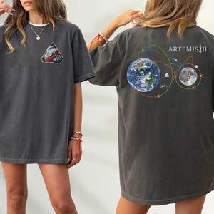 Puede incluir: Camiseta gris oscuro con cuello blanco. La parte delantera presenta un parche triangular con un gráfico de temática espacial. La parte trasera muestra un gráfico de la Tierra, la Luna y líneas de conexión, con el texto "ARTEMIS II".