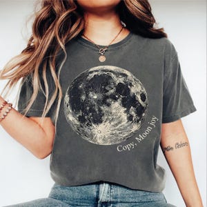 Puede incluir: Camiseta gris oscuro con una ilustración detallada en blanco y negro de la luna. El texto "Copy, Moon joy" está impreso en la camiseta. Un estilo casual y cómodo.