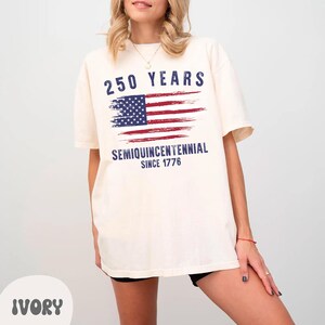May include: Ivory-colored t-shirt with a distressed American flag graphic. The shirt features the text "250 YEARS" above the flag and "SEMIQUINCENTENNIAL SINCE 1776" below. The shirt is a relaxed fit.